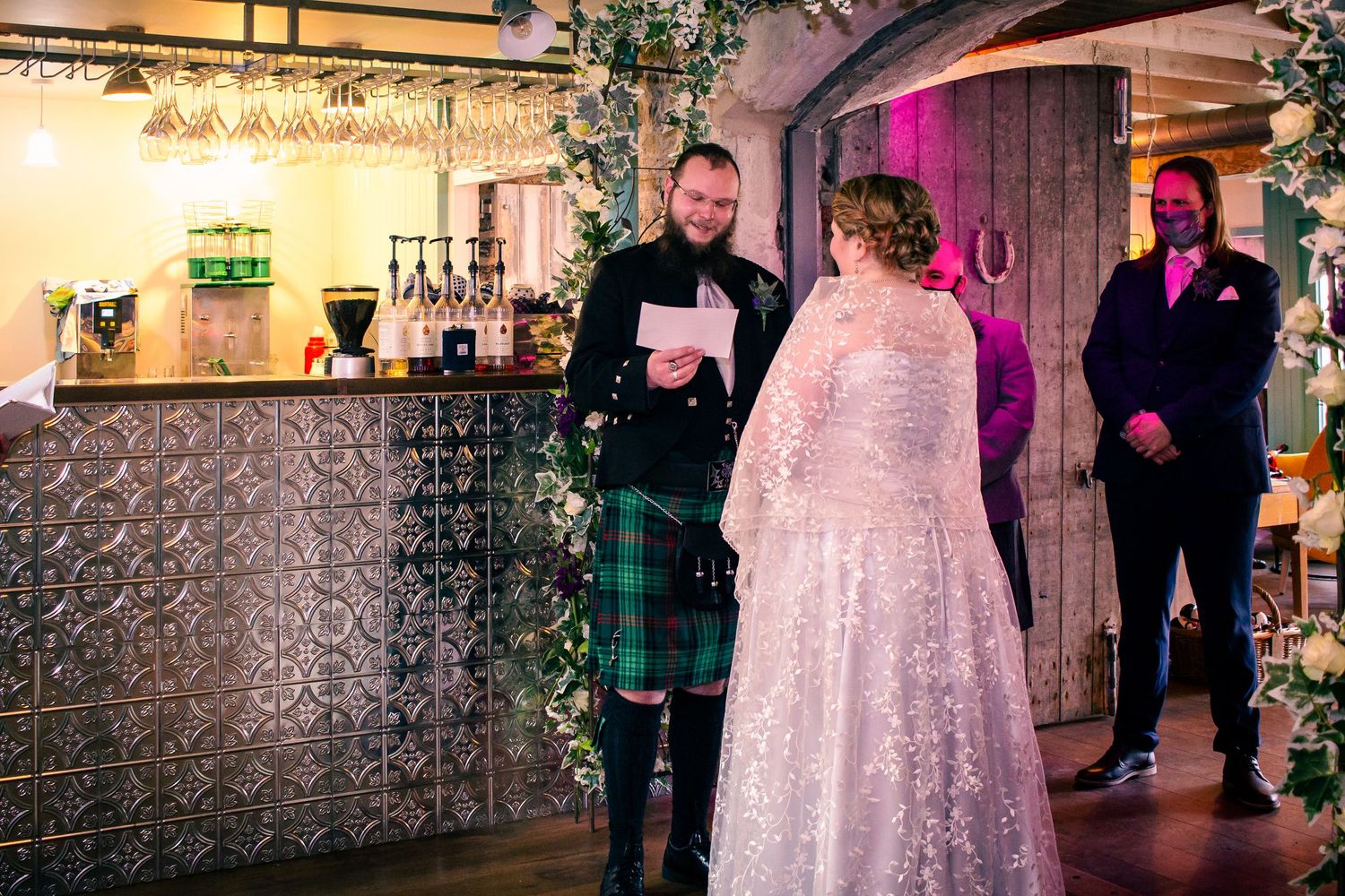 Groom in tartan kilt reads vows to bride in lace wedding dress at elegant indoor wedding ceremony at the storehouse, kirkwall.