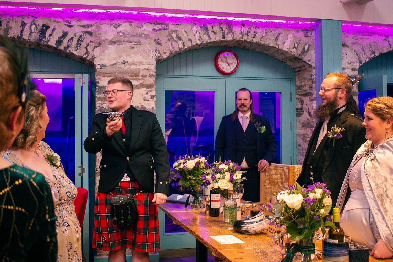 Wedding guests in Scottish kilts gather around a decorated table in a stone venue with purple lighting at the storehouse, kirkwall.