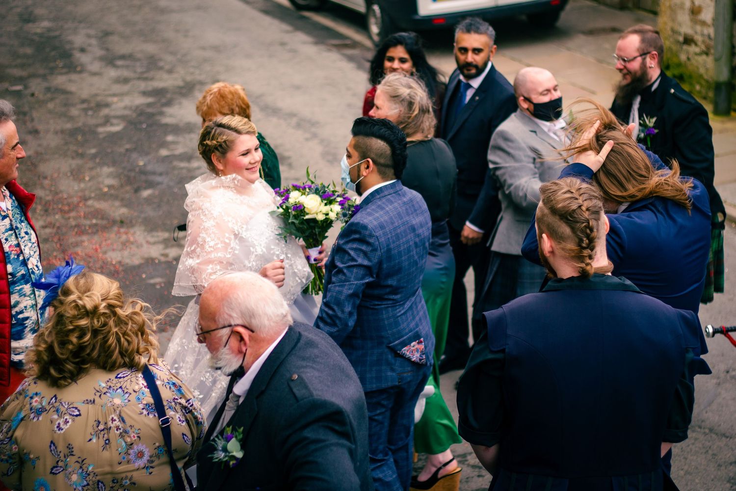 Bride in lace dress holding bouquet greeted by wedding guests outdoors at elegant ceremony.