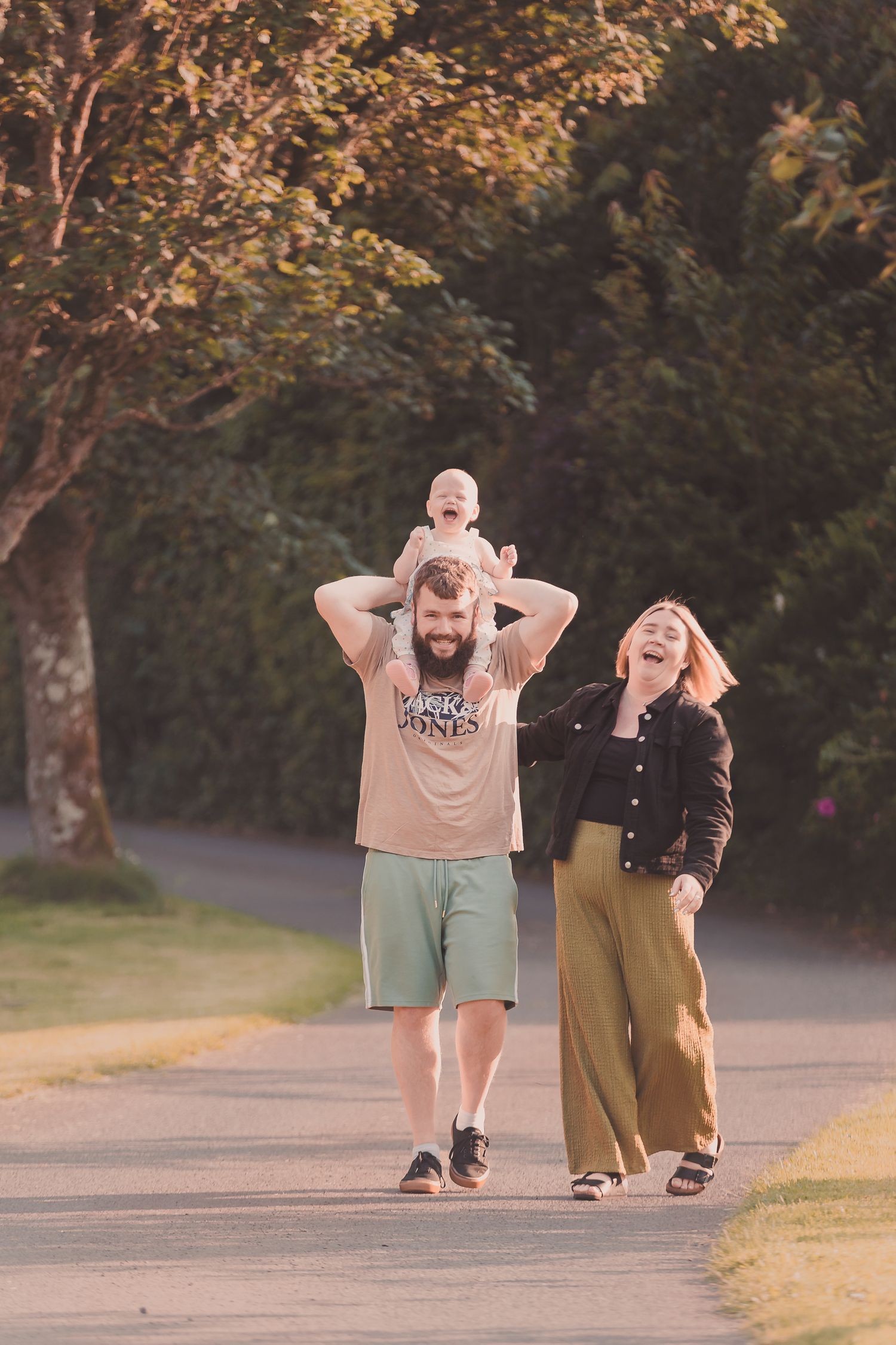 Joyful family walking on a tree-lined path with baby on fathers shoulders at golden hour by Orkney Family Photographer