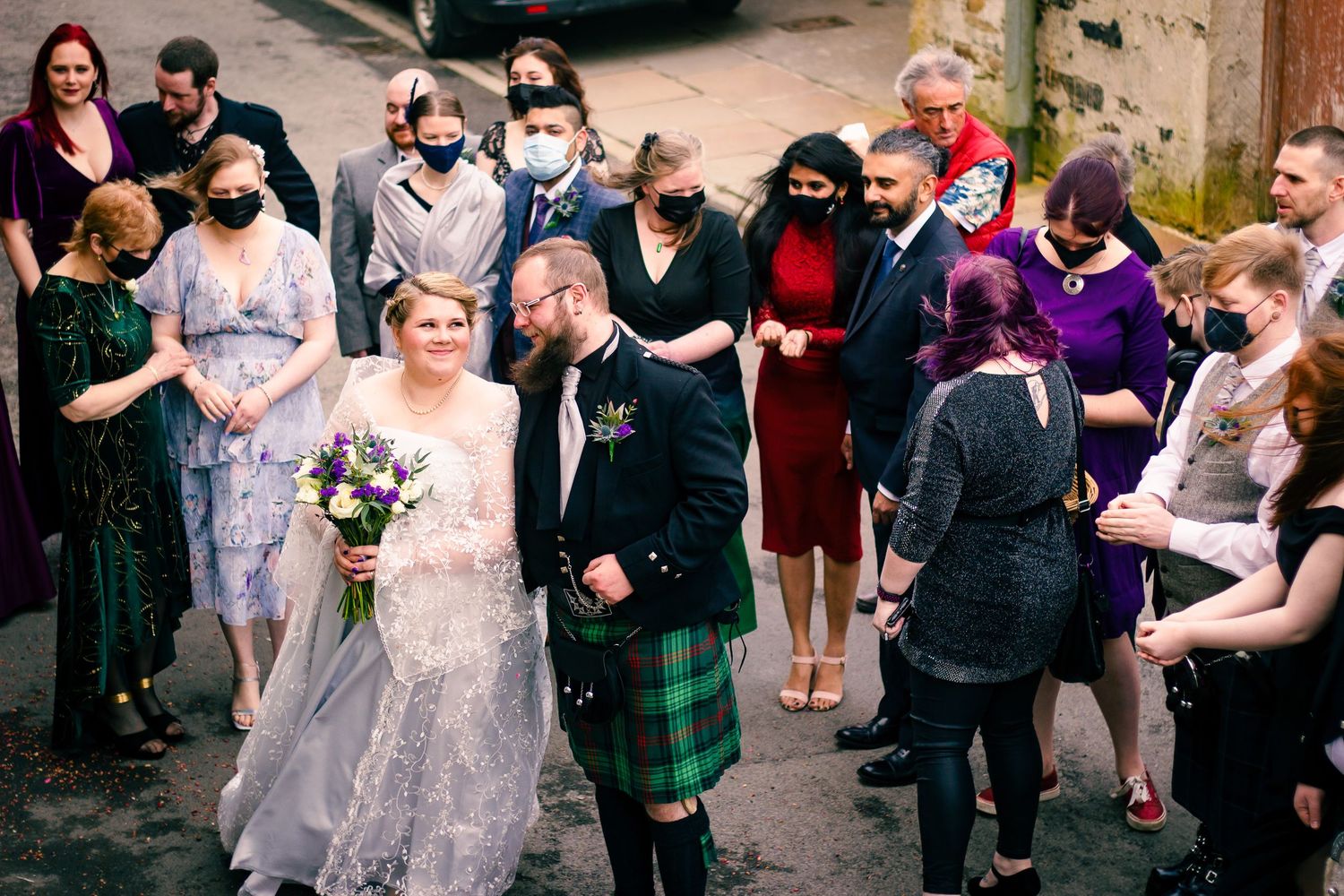 Bride and groom in Scottish kilt walk with masked wedding guests on cobblestone street, bride holding purple bouquet.