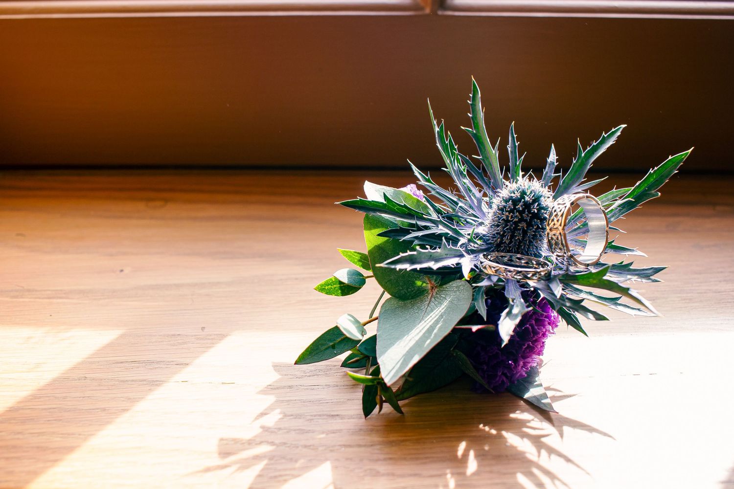 Thistle and greenery boutonniere with silver ring detail resting on a wooden floor in warm sunlight.