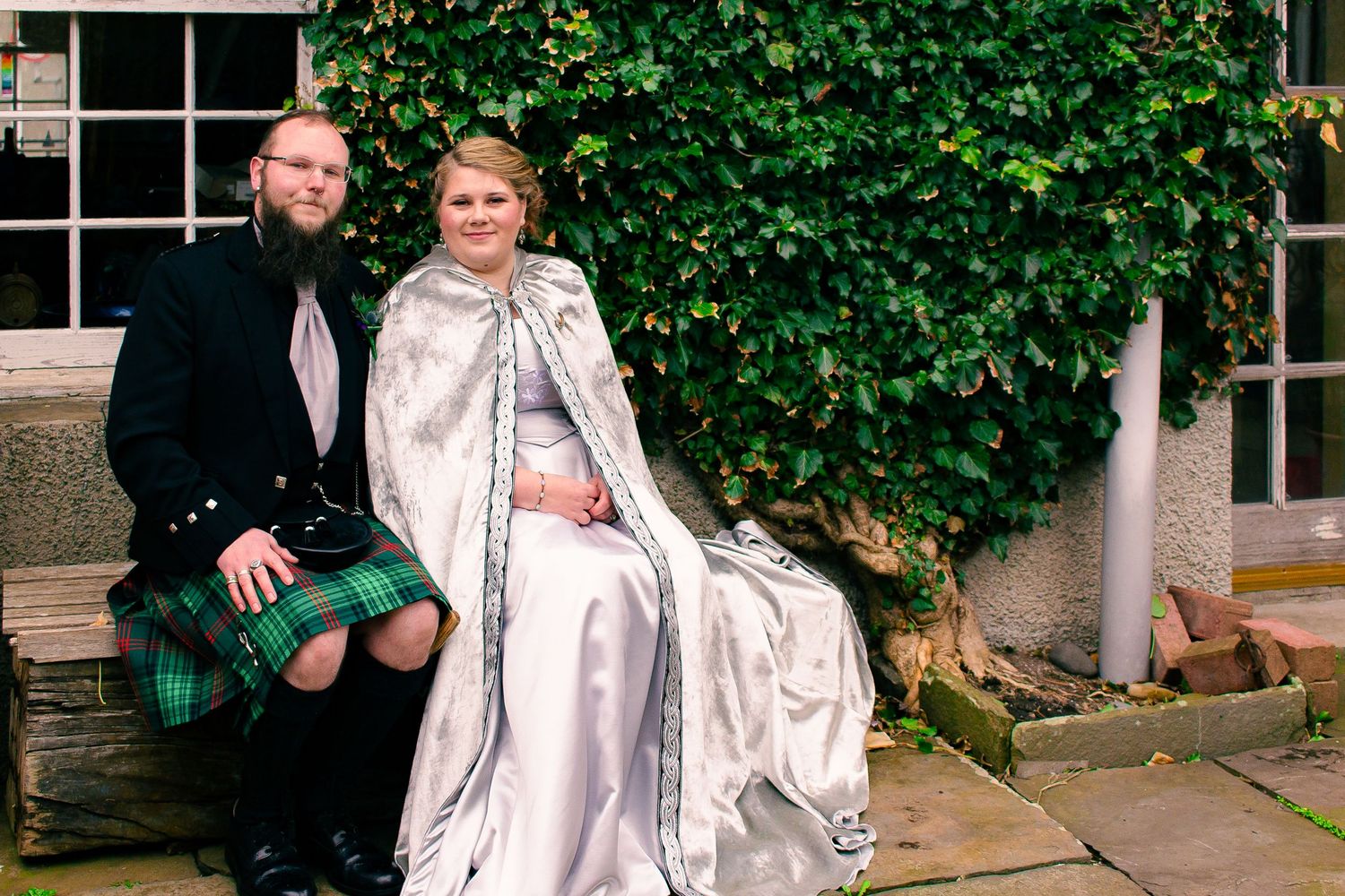 Couple in Scottish wedding attire, man in green tartan kilt and woman in silver cape gown, seated outdoors.