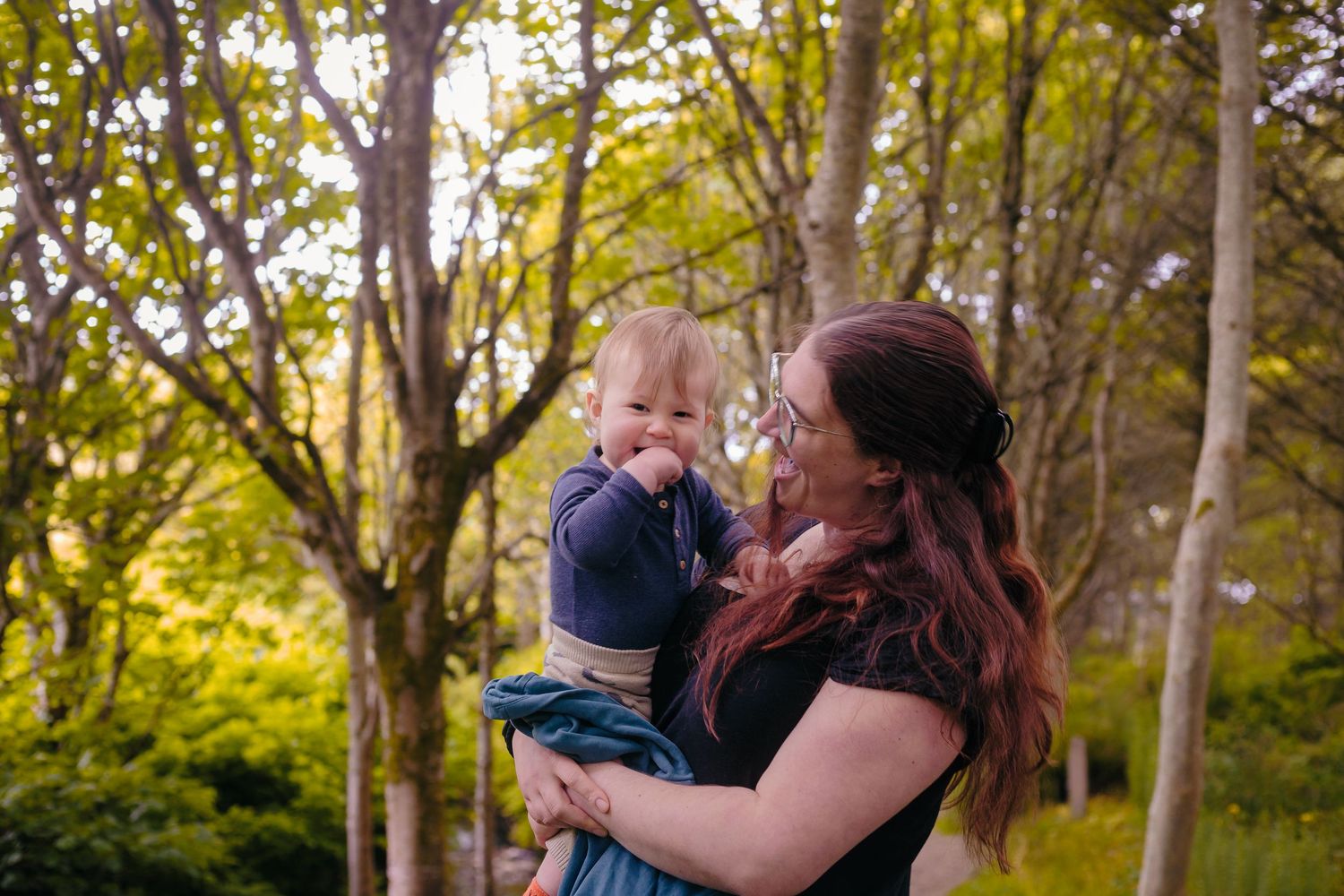 Smiling mother holding her laughing baby boy among tall green trees in sunlit Happy Valley,  by Orkney Family Photographer