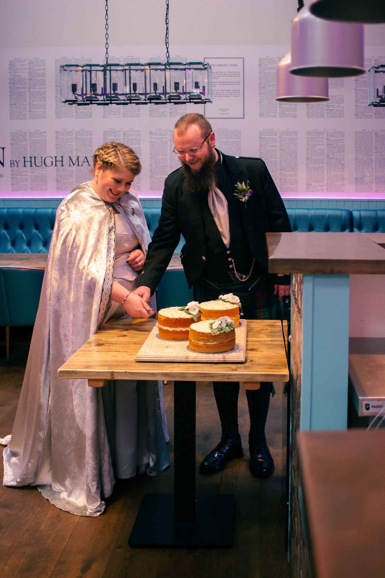Couple in wedding attire cutting a layered cake on a wooden table in a decorated venue at the storehouse, kirkwall.