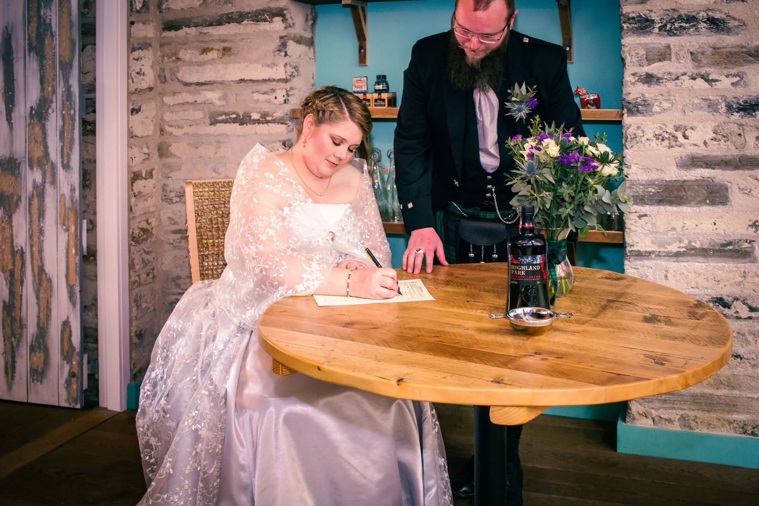 Bride in lace gown signing wedding register at wooden table with groom in kilt standing beside her at the storehouse, kirkwall.