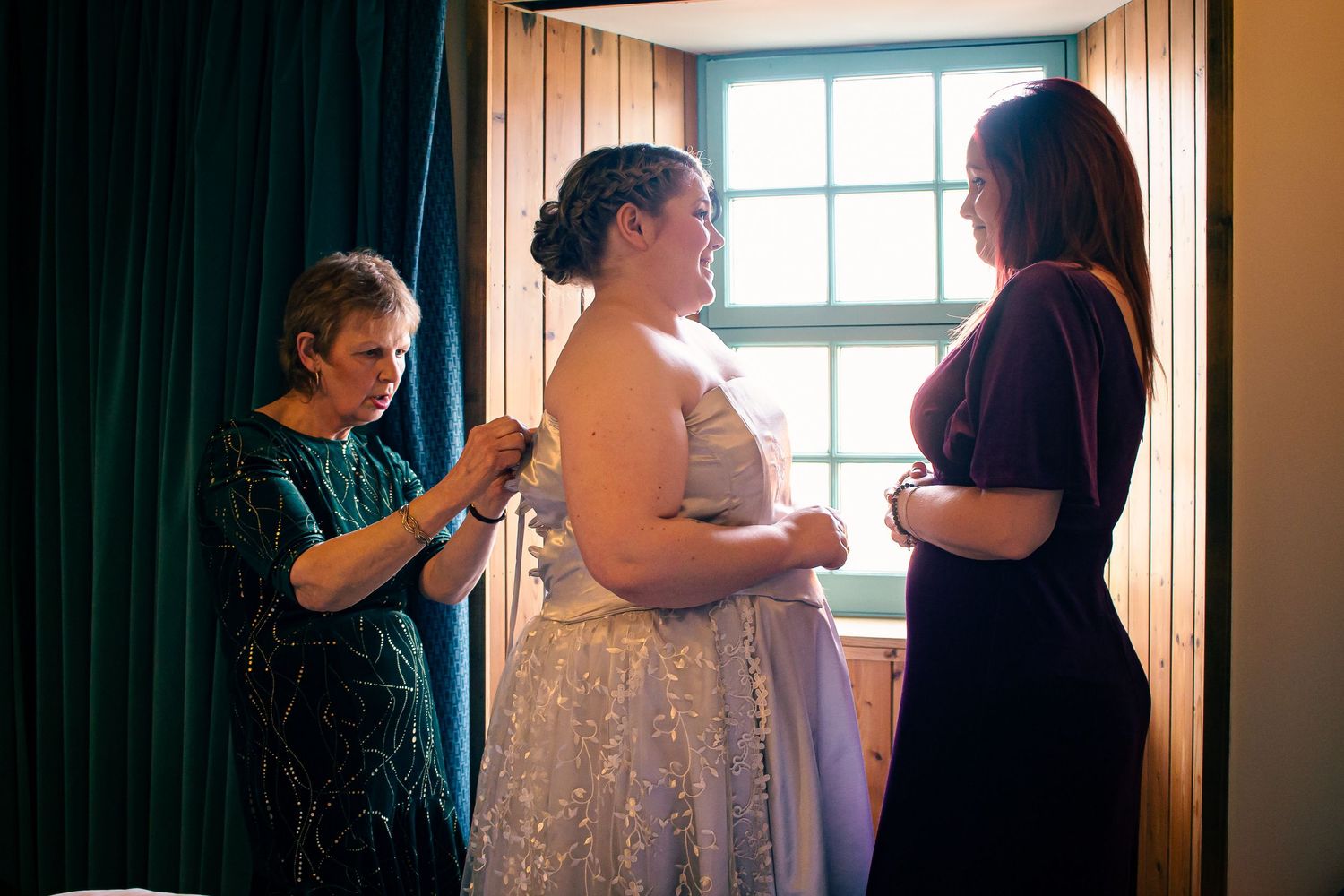 Bride in sparkly wedding gown being helped by two women in a warmly lit room in orkney.