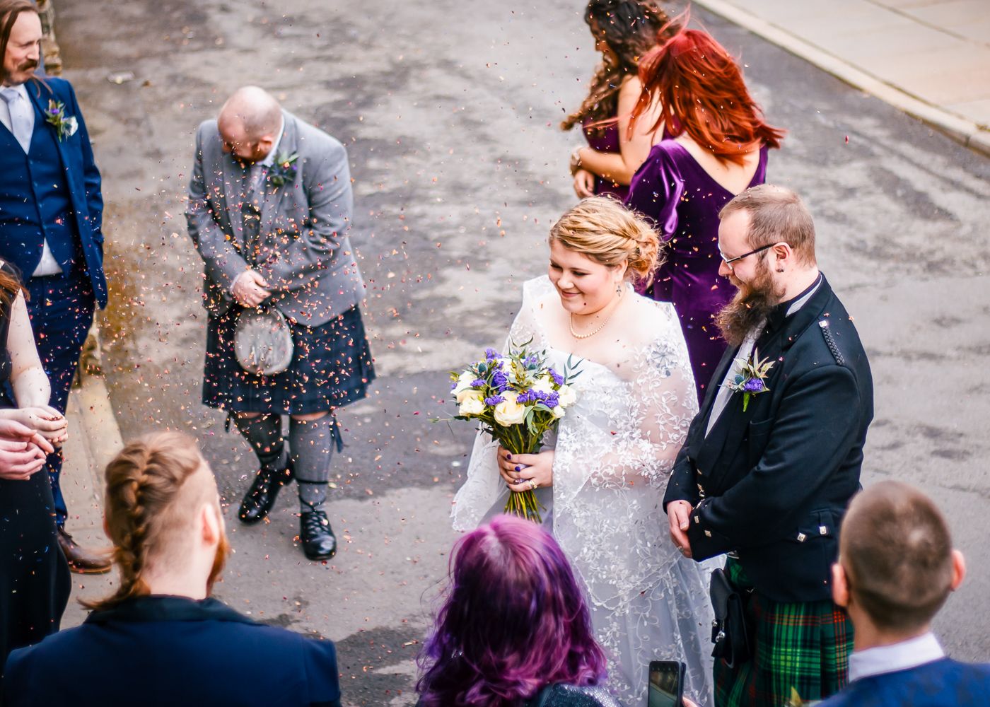 Wedding guests toss confetti over a smiling bride holding wildflowers and groom in a tartan kilt in Kirkwall taken by Orkney Photographer