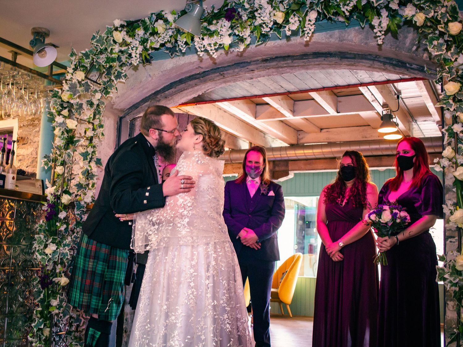Couple shares first kiss at wedding altar decorated with flowers, groom in kilt, bridesmaids in purple dresses nearby at the storehouse.