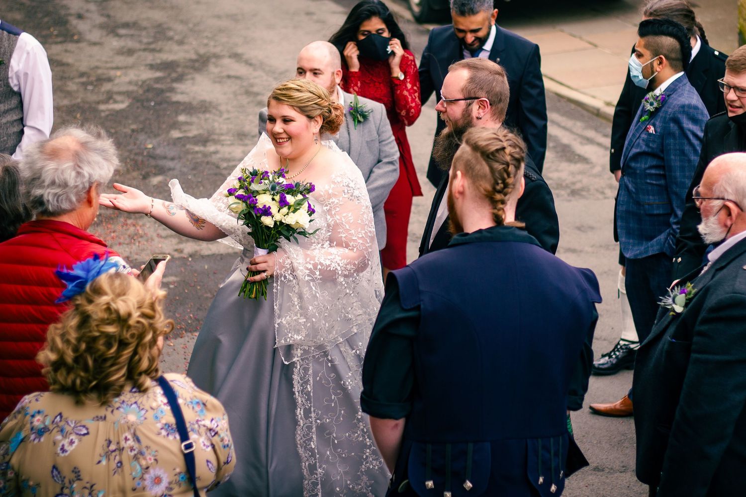 Bride in lace wedding gown holds purple bouquet greeting guests outdoors at a wedding celebration.