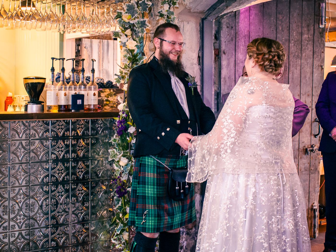 A bearded groom in a dark tartan kilt holds hands with his bride in a lace gown at the altar in Kirkwall restaurant taken by Orkney Wedding