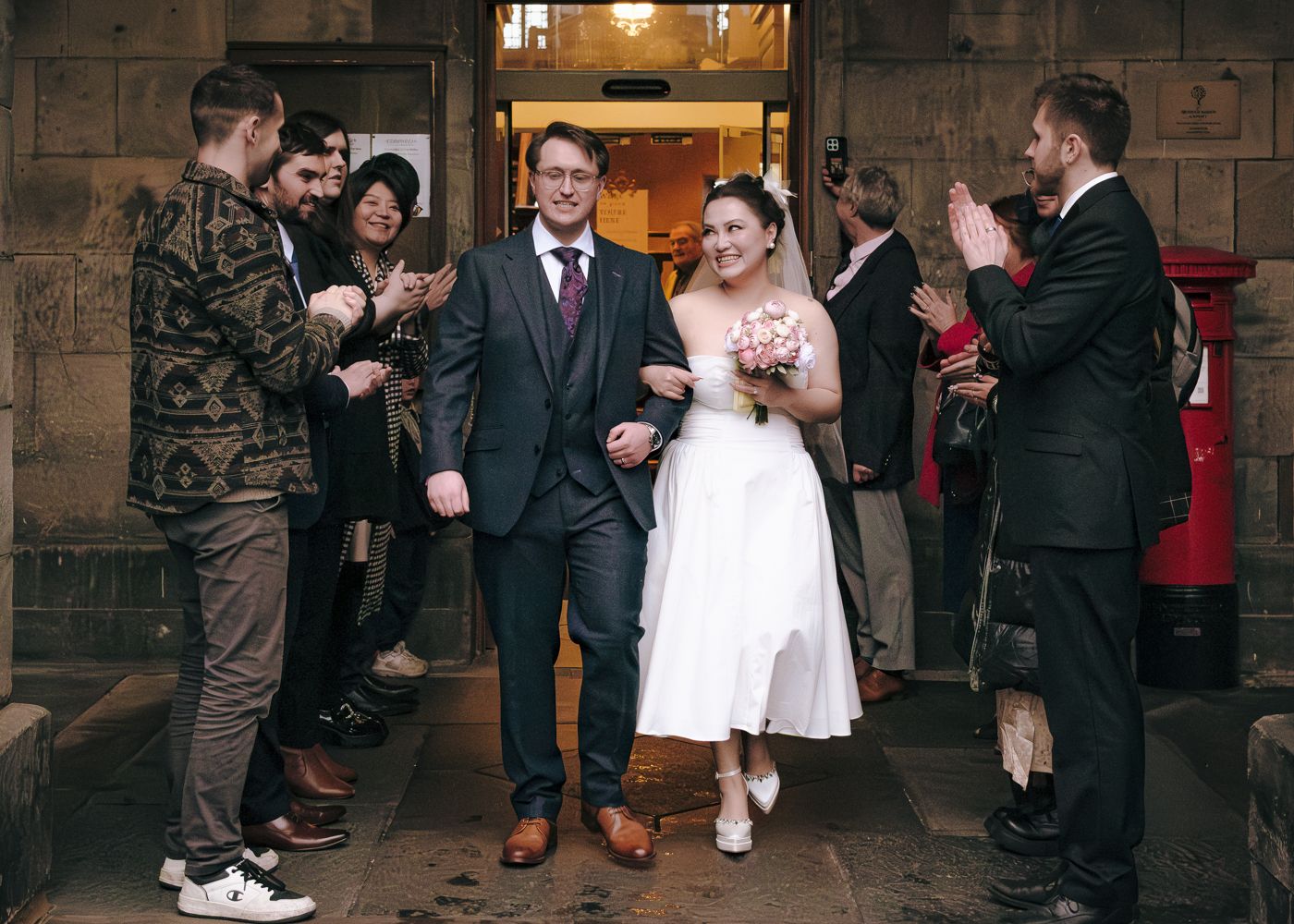 Newlyweds walk through cheering guests outside a historic stone building after their wedding ceremony in Edinburgh