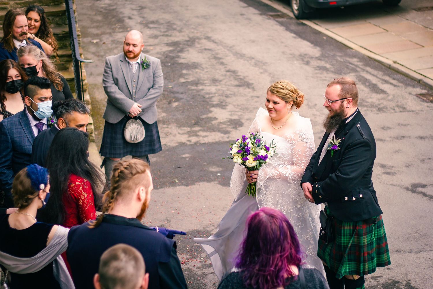 Bride and groom in Scottish kilts share a joyful moment outdoors surrounded by wedding guests holding a colorful bouquet.