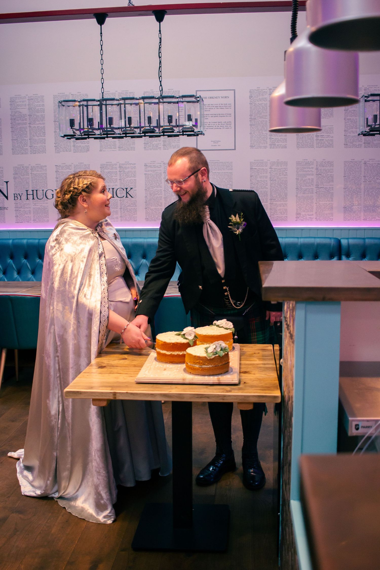 Bride in white cape and groom in kilt cutting wedding cake at rustic wooden table at the storehouse, kirkwall.