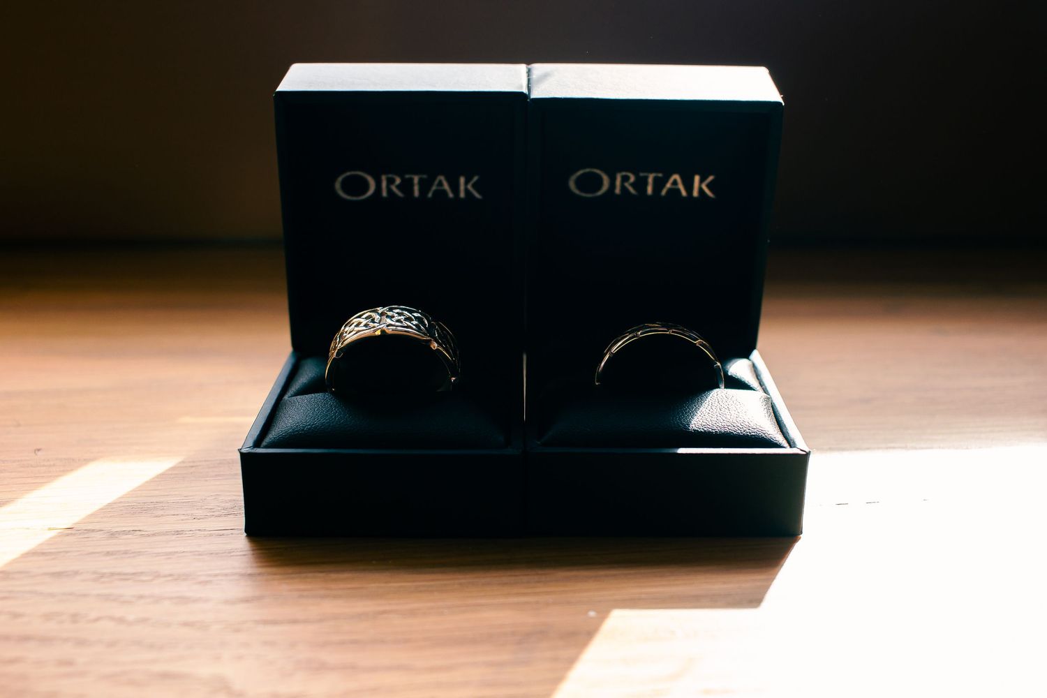 Two Ortak wedding rings displayed in black gift boxes on a wooden surface.