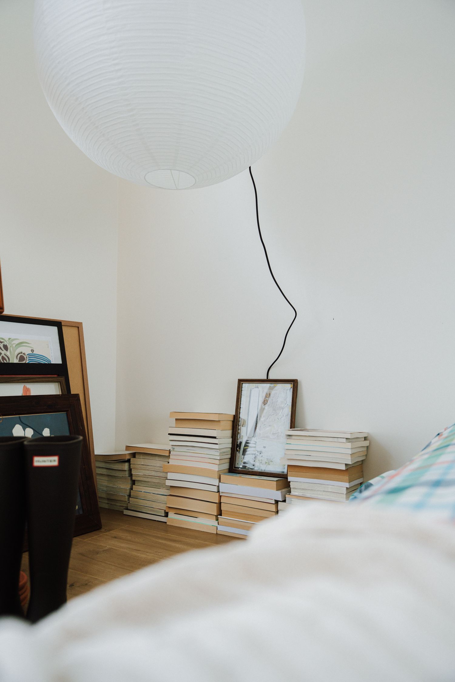 Stack of books with framed artwork beside a white paper lantern lamp in a minimalist bedroom.