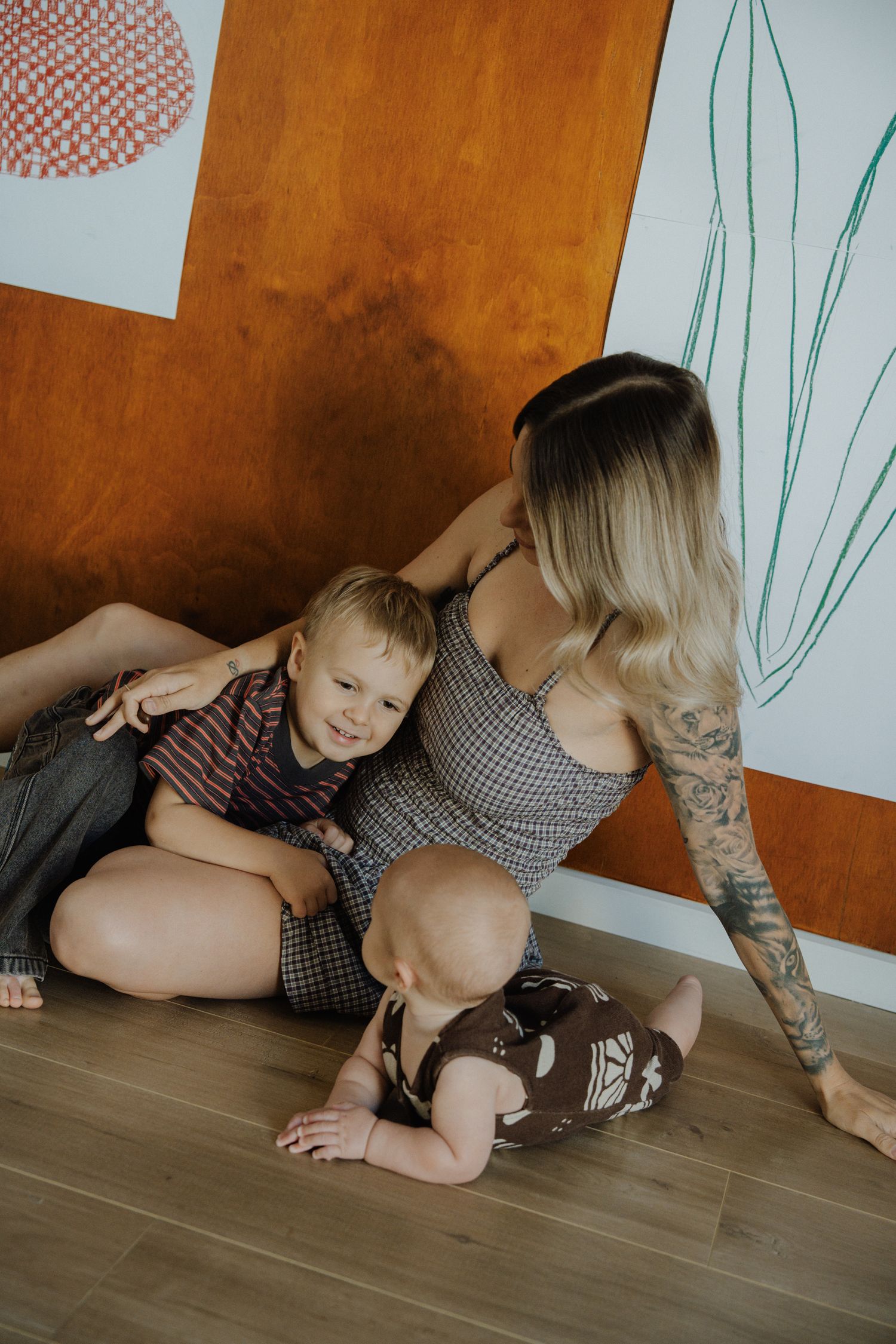 Mother with tattoos sitting on floor playing with two young children against colorful wall art.