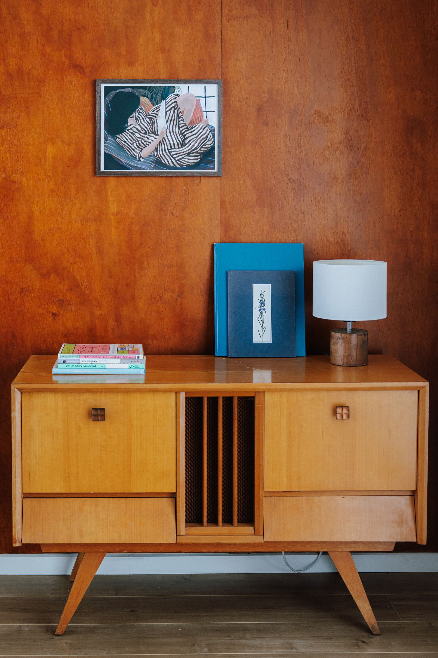 Mid-century modern wooden sideboard with lamp and artwork against warm brown paneled wall.
