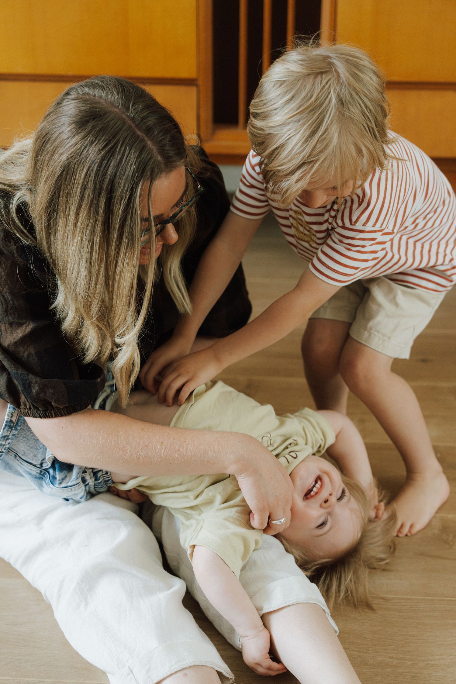 Mother and young boy playing with laughing baby on wooden floor at home.