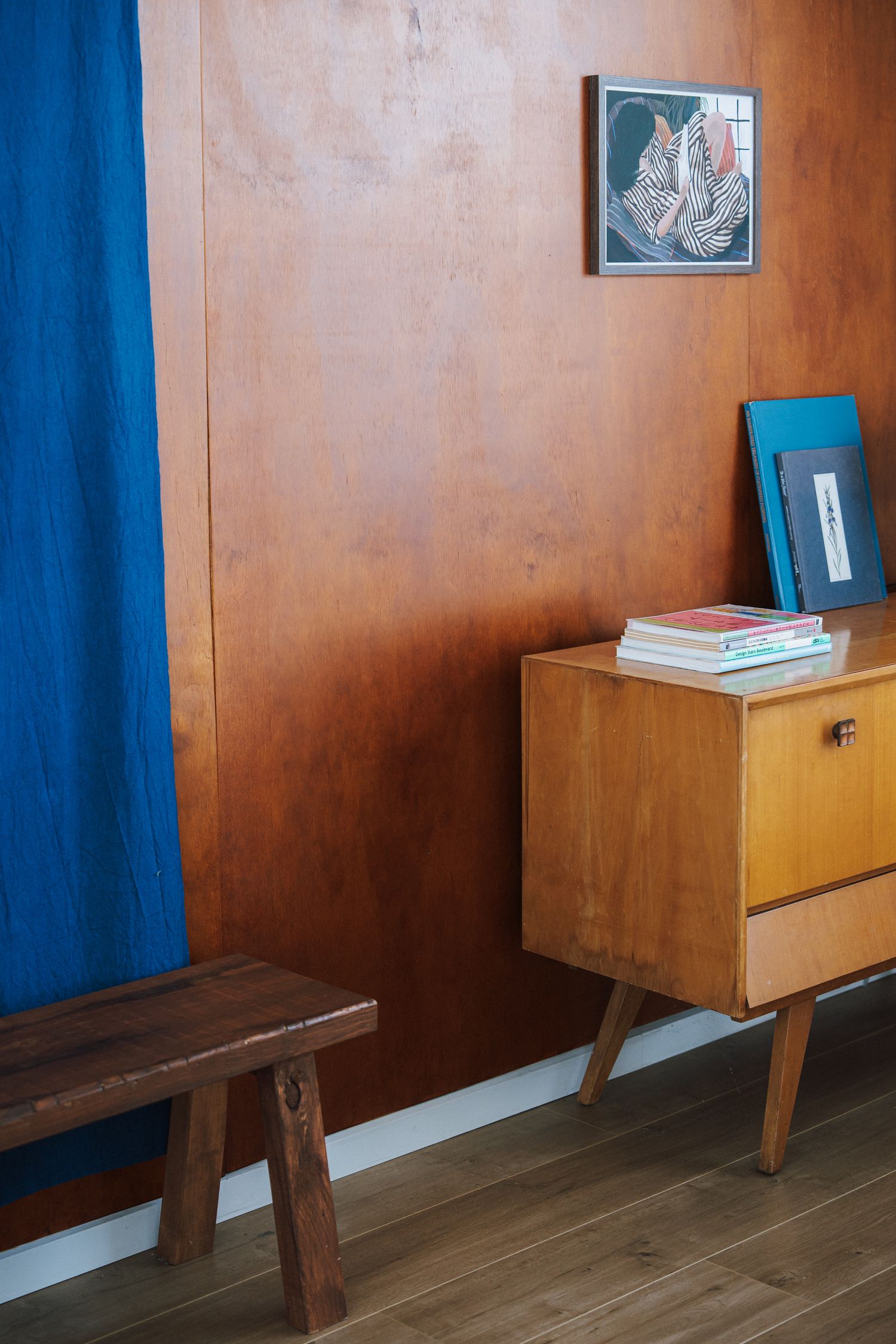 Vintage wooden dresser with books and artwork against warm brown wood-paneled wall with blue curtain.