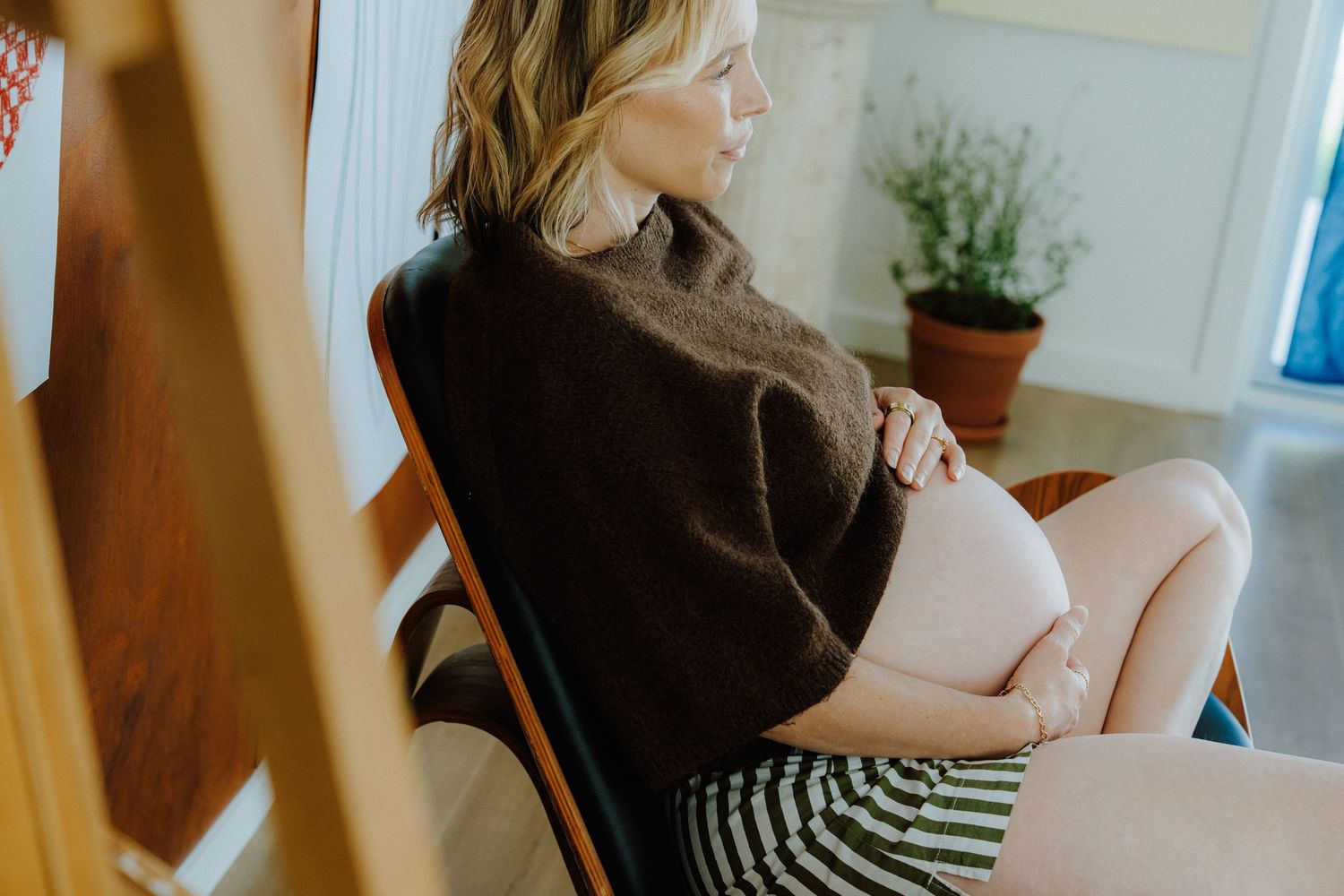 Pregnant woman in brown sweater sitting in wooden chair cradling her belly by a bright window.
