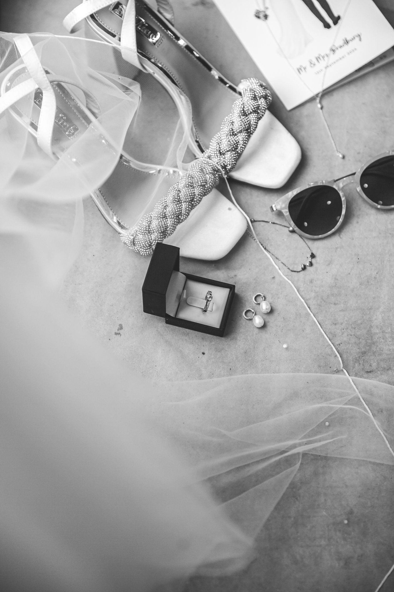 Black and white flat lay of bridal accessories including heels, veil, ring box, sunglasses, and pearl necklace.