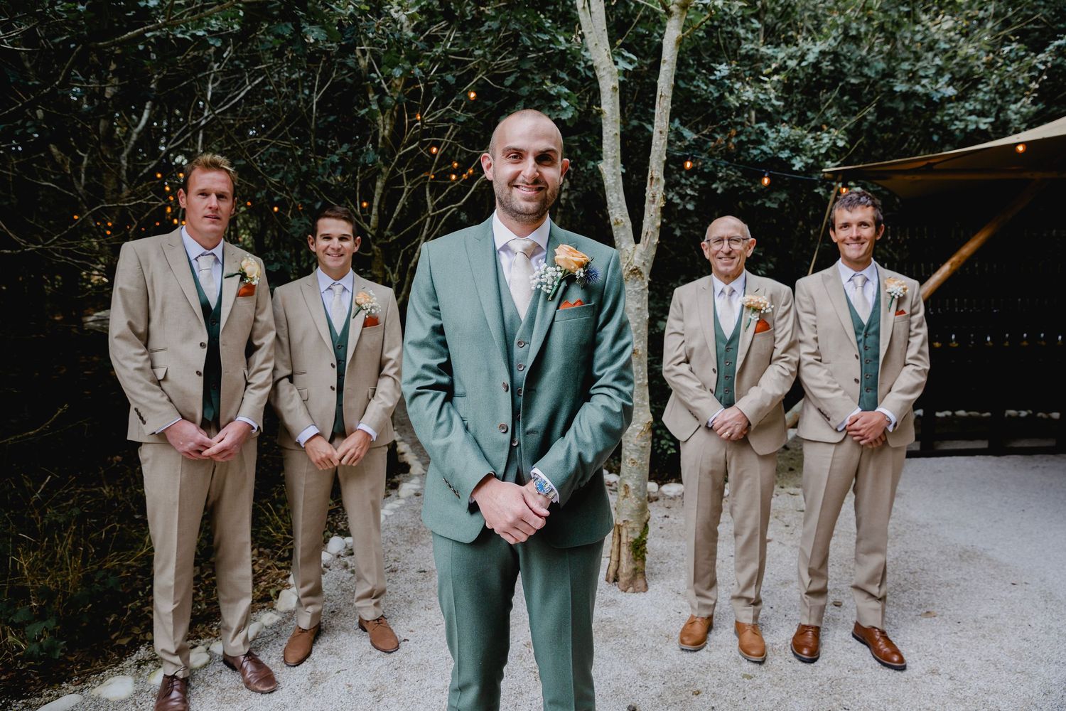 Groom in green suit stands before four groomsmen in tan suits at outdoor wedding venue.