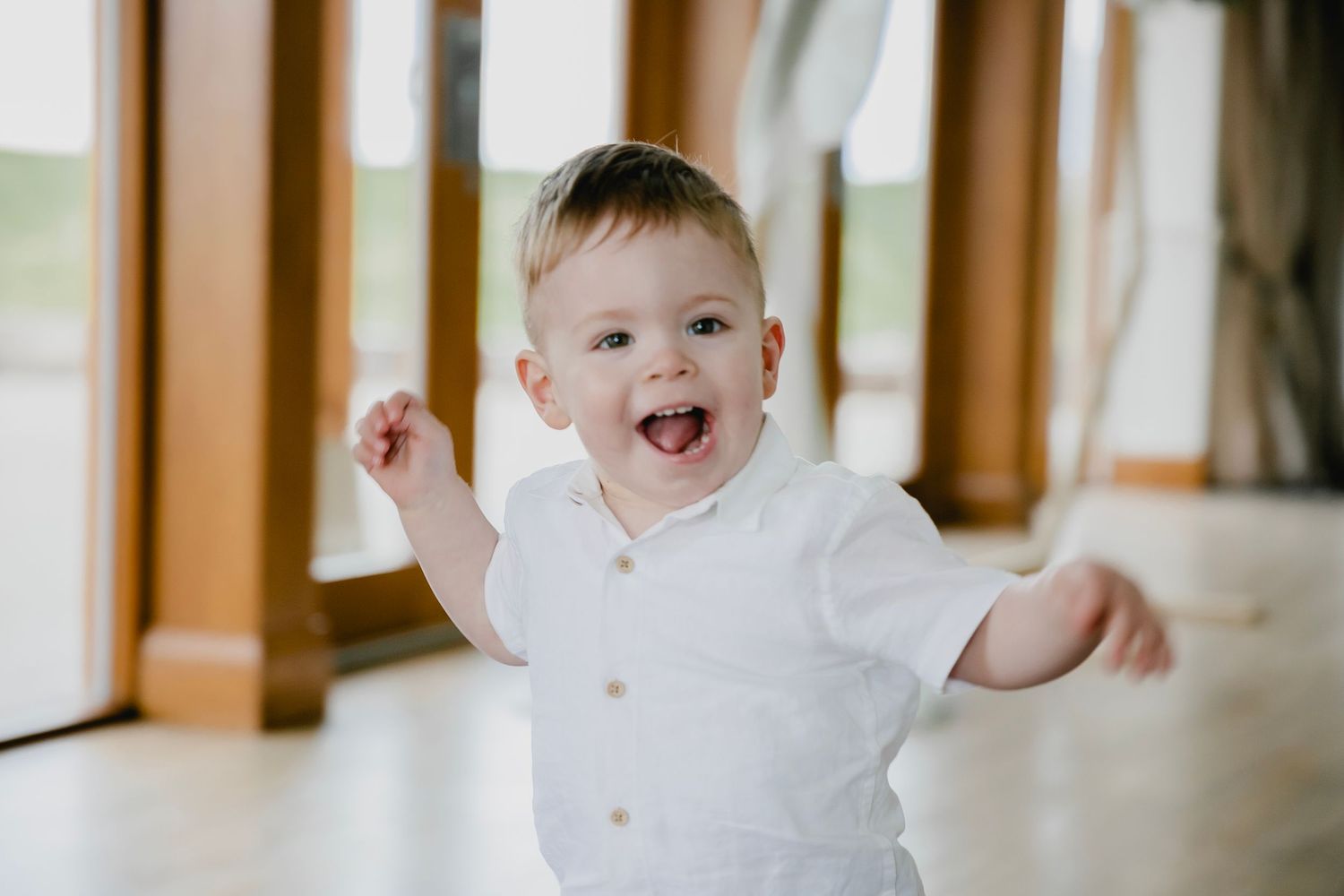 Adorable smiling baby boy in white outfit with arms outstretched at wedding venue.