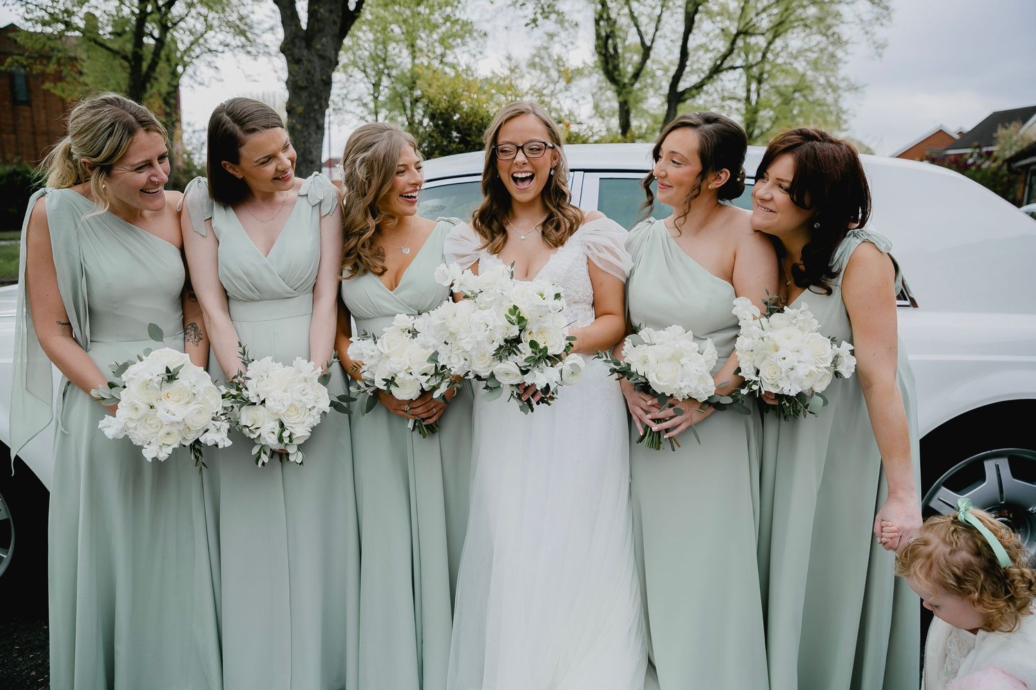 Bride with bridesmaids in sage green dresses holding white bouquets beside a white car.
