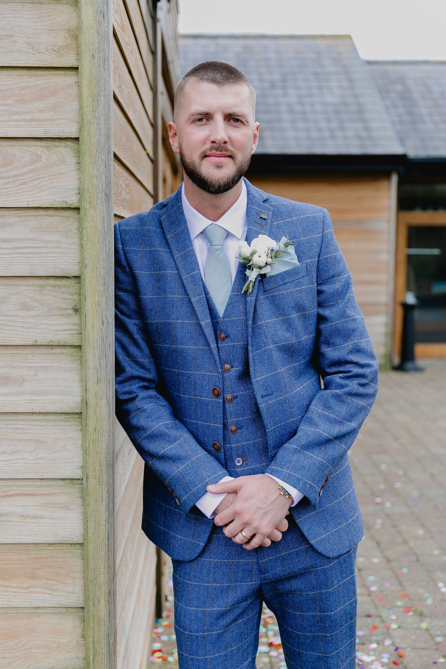 Groom in blue check three-piece suit with white boutonniere leaning against wooden barn wall.