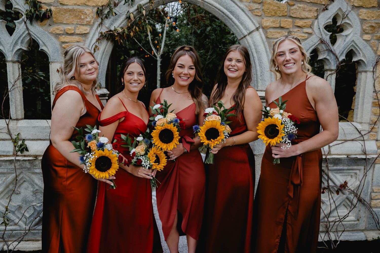 Five bridesmaids in rust and red dresses holding sunflower bouquets pose before gothic stone archway.