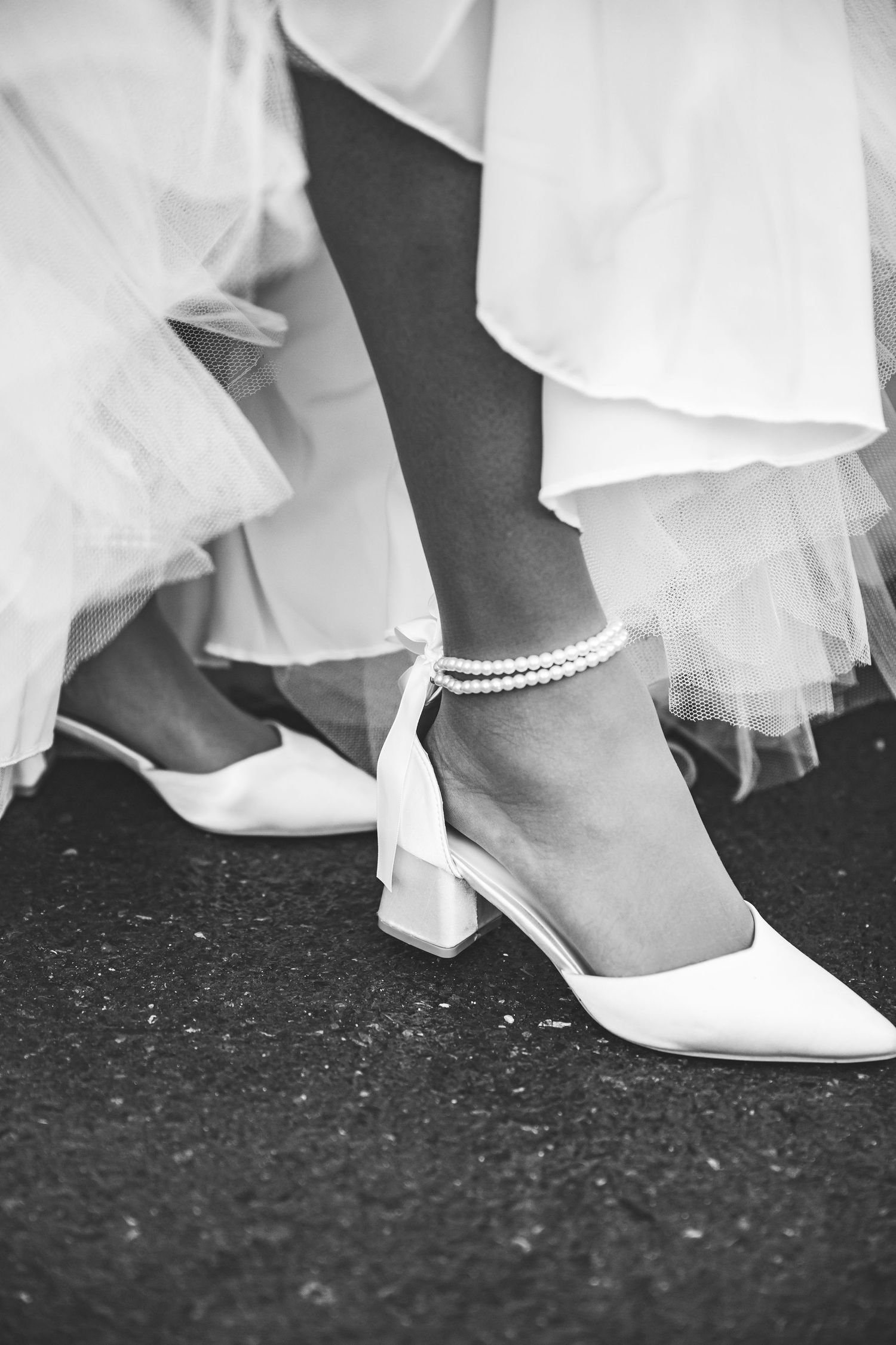 Black and white close-up of bride wearing white block-heel shoes with delicate pearl ankle bracelet and tulle gown.