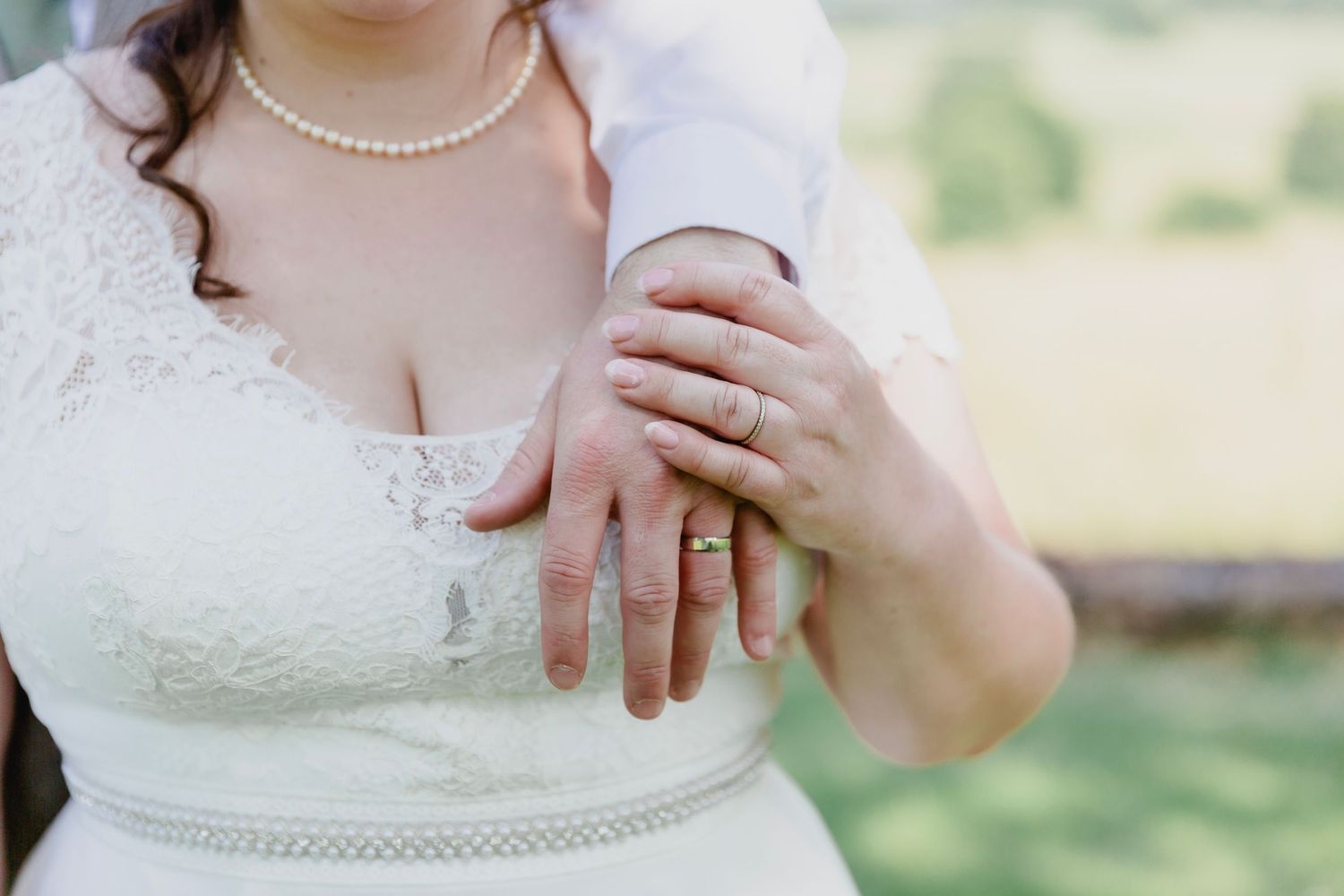 Bride and groom showing wedding rings on hands, wearing white lace dress outdoors.