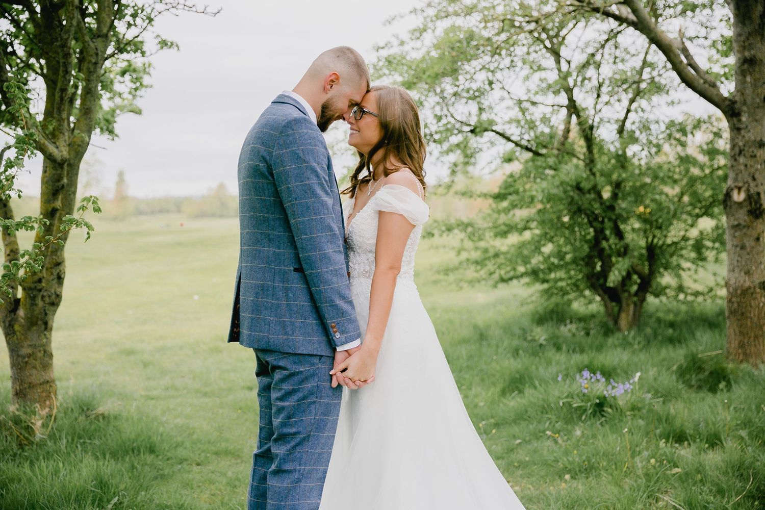 Bride and groom holding hands touching foreheads in lush green countryside wedding setting.