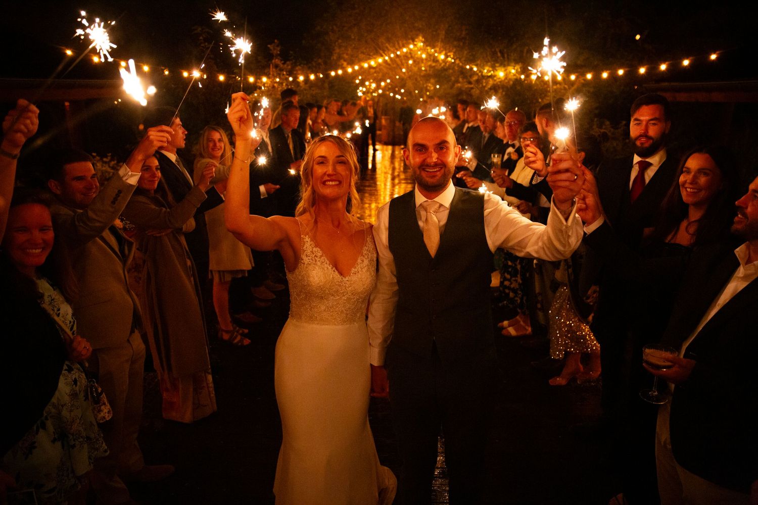 Bride and groom walk through sparkler send-off surrounded by cheering wedding guests at night.