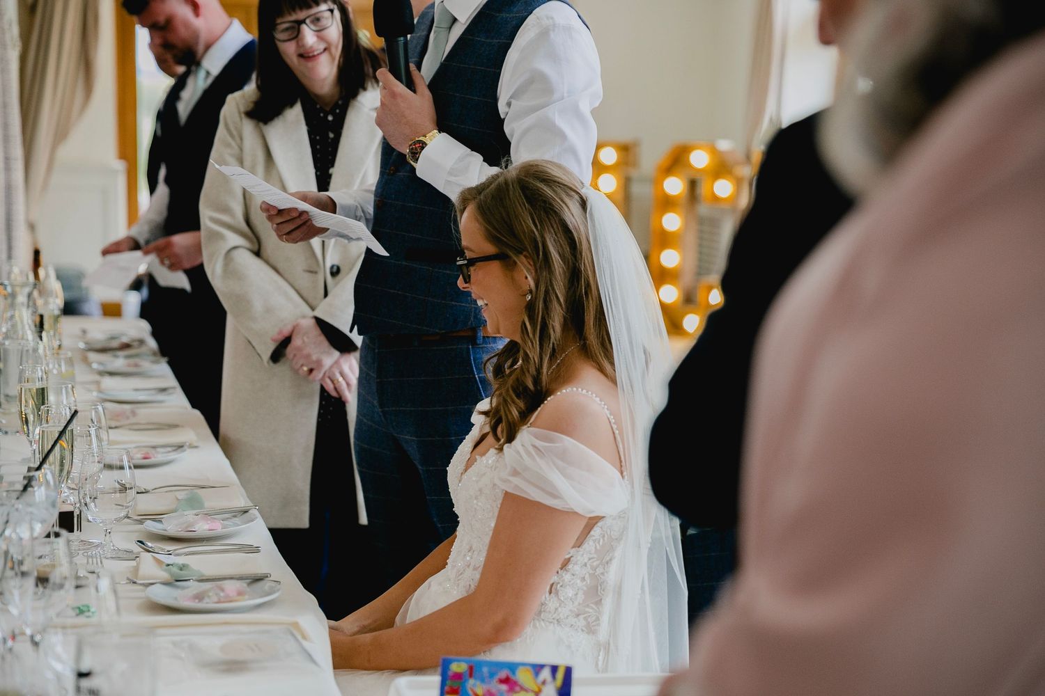 Bride in white off-shoulder gown smiling at elegantly decorated wedding reception table.