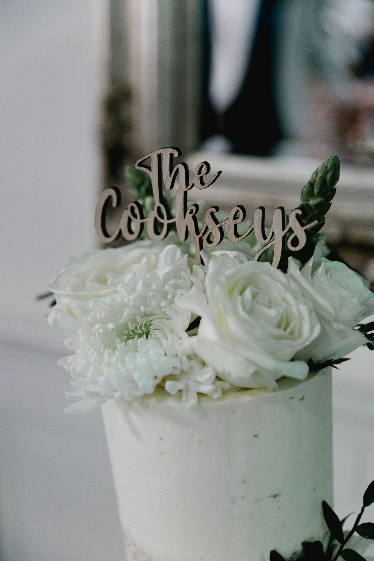 White floral wedding cake topper reading The Cookseys surrounded by white roses and chrysanthemums.