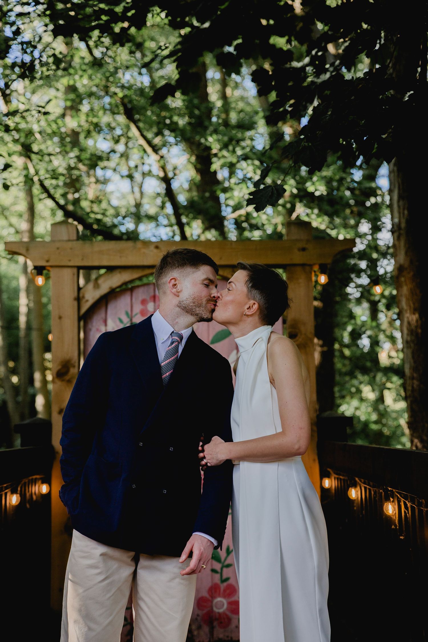 A couple shares a romantic kiss under a wooden arch surrounded by lush green trees outdoors.
