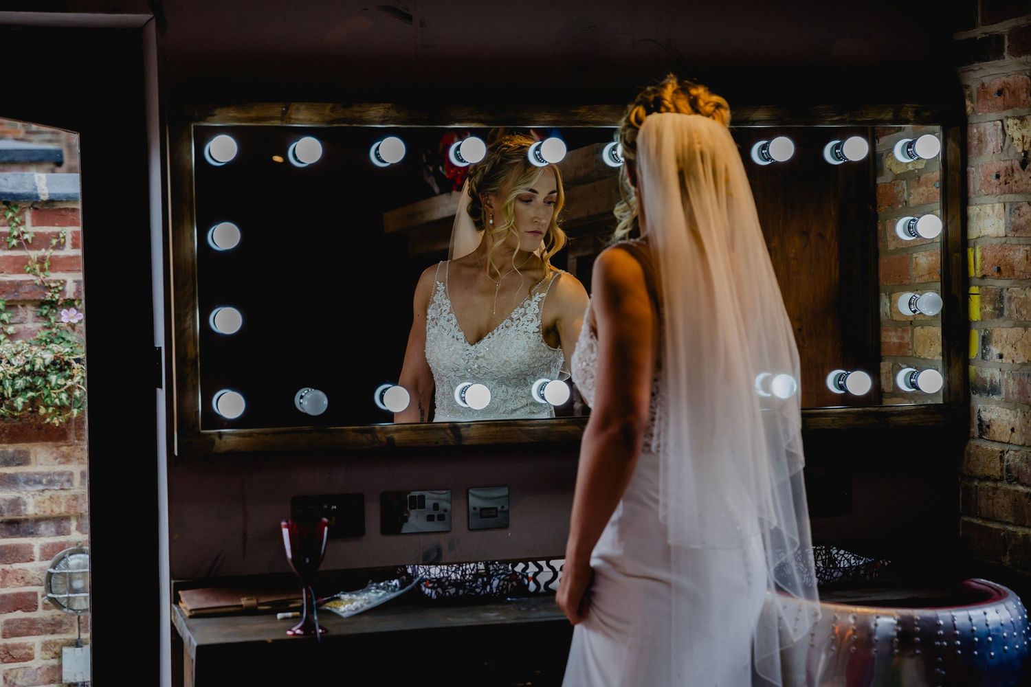 Bride in lace wedding dress viewing her reflection in a Hollywood mirror with bright bulbs.