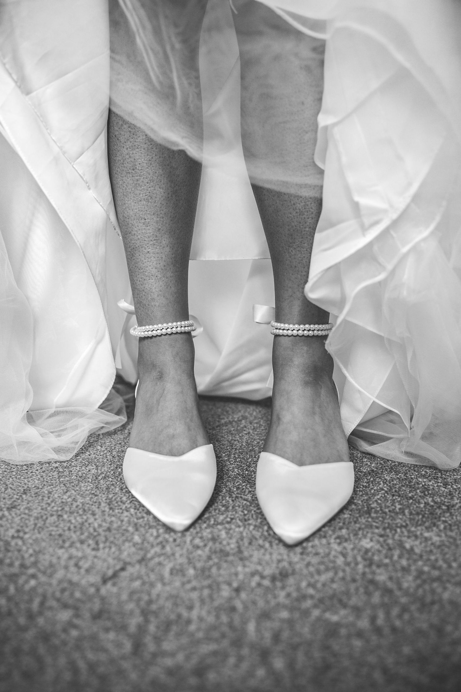 Black and white close-up of bride wearing white satin pointed-toe heels with pearl ankle straps.