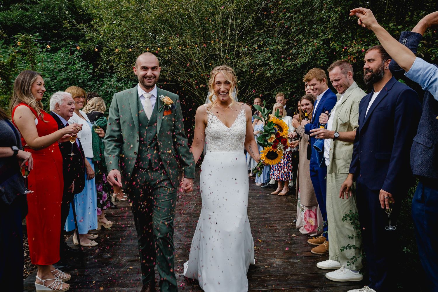Newlyweds walk through confetti shower lined by cheering guests on garden path after ceremony.