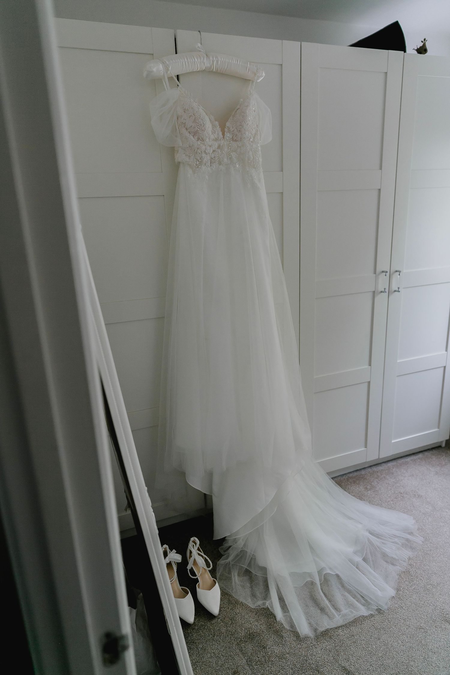Elegant white off-shoulder lace and tulle wedding dress hanging with bridal heels below.