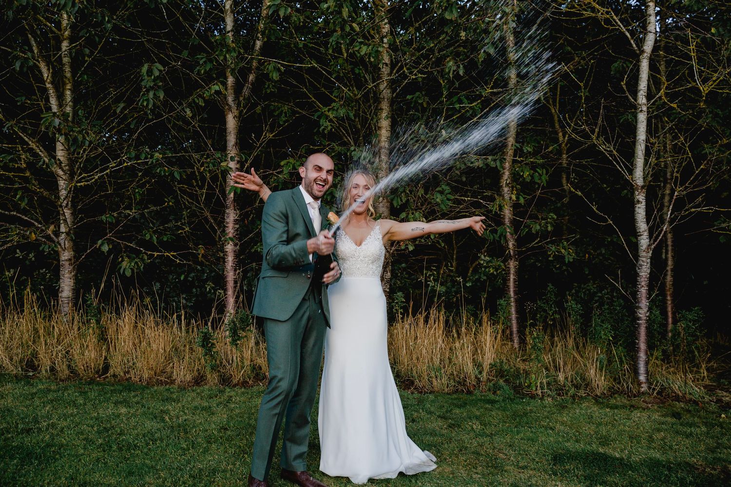 Bride and groom celebrating with champagne spray in front of woodland trees at outdoor wedding.