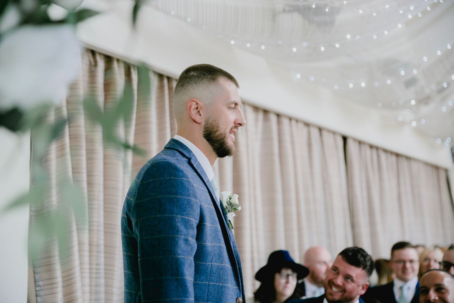 Groom in blue checked suit smiling at wedding ceremony with guests seated behind.
