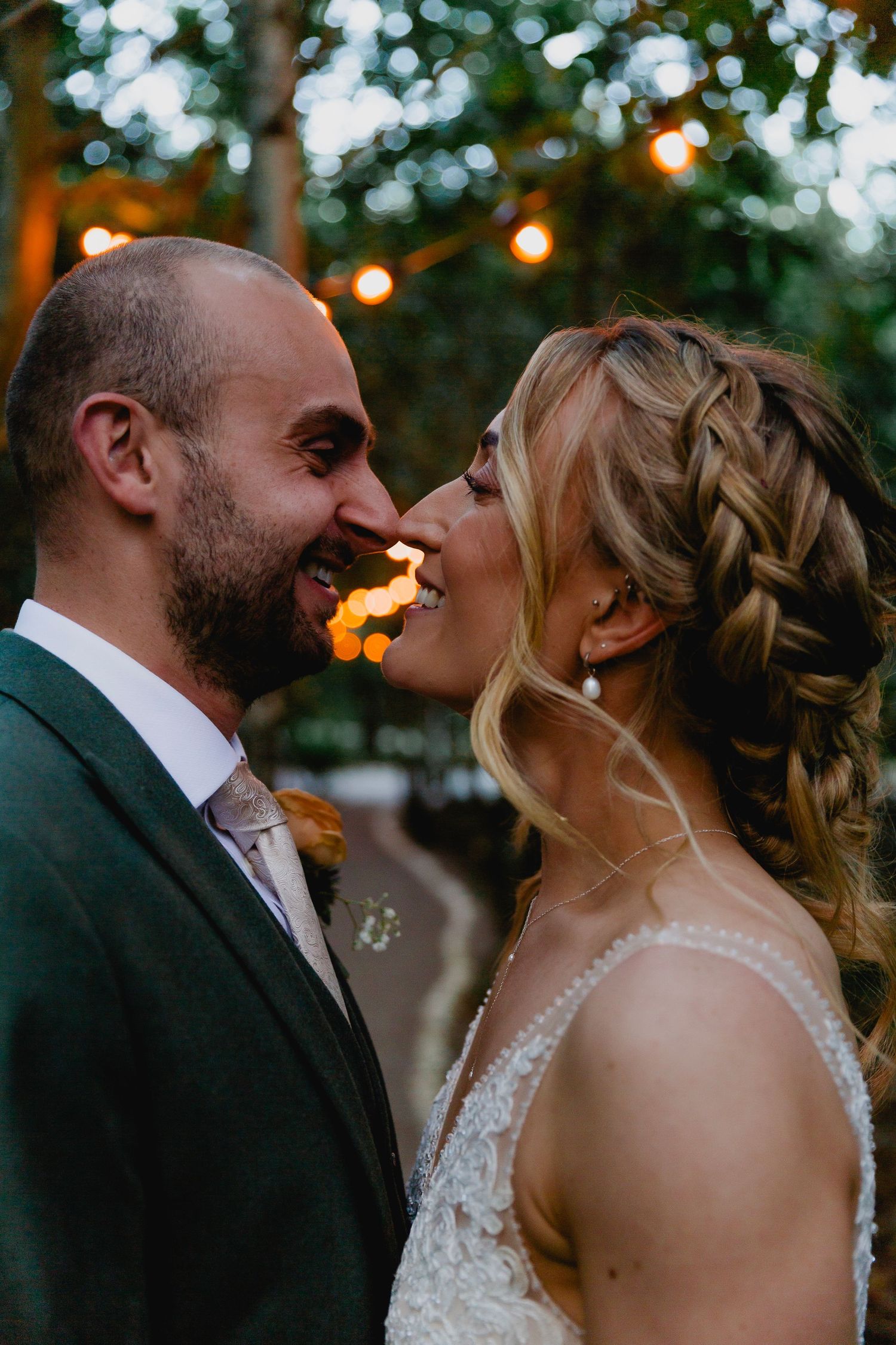 Smiling bride and groom almost kissing under warm fairy lights at romantic outdoor evening wedding.