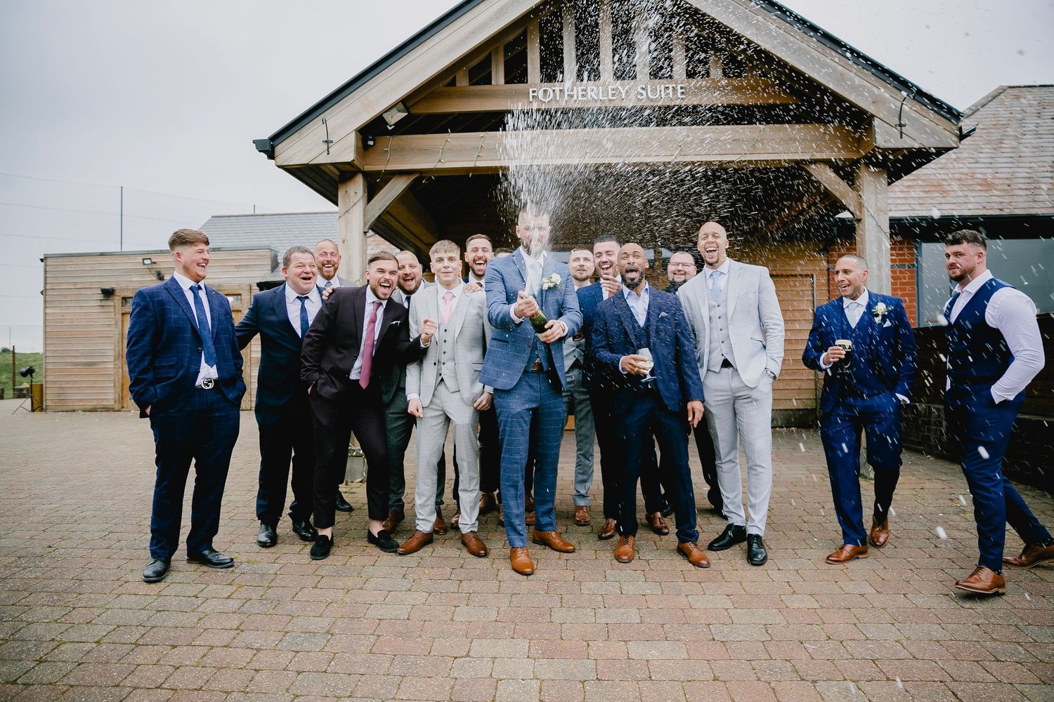 Groom and groomsmen celebrating with champagne spray outside rustic barn wedding venue.