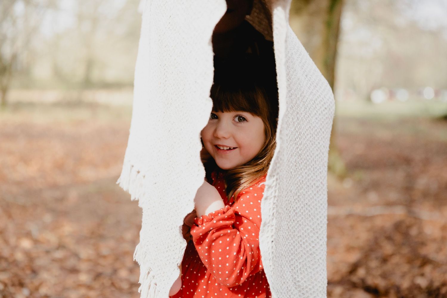 A young girl in a red polka dot dress peeks through a white knit blanket outdoors.