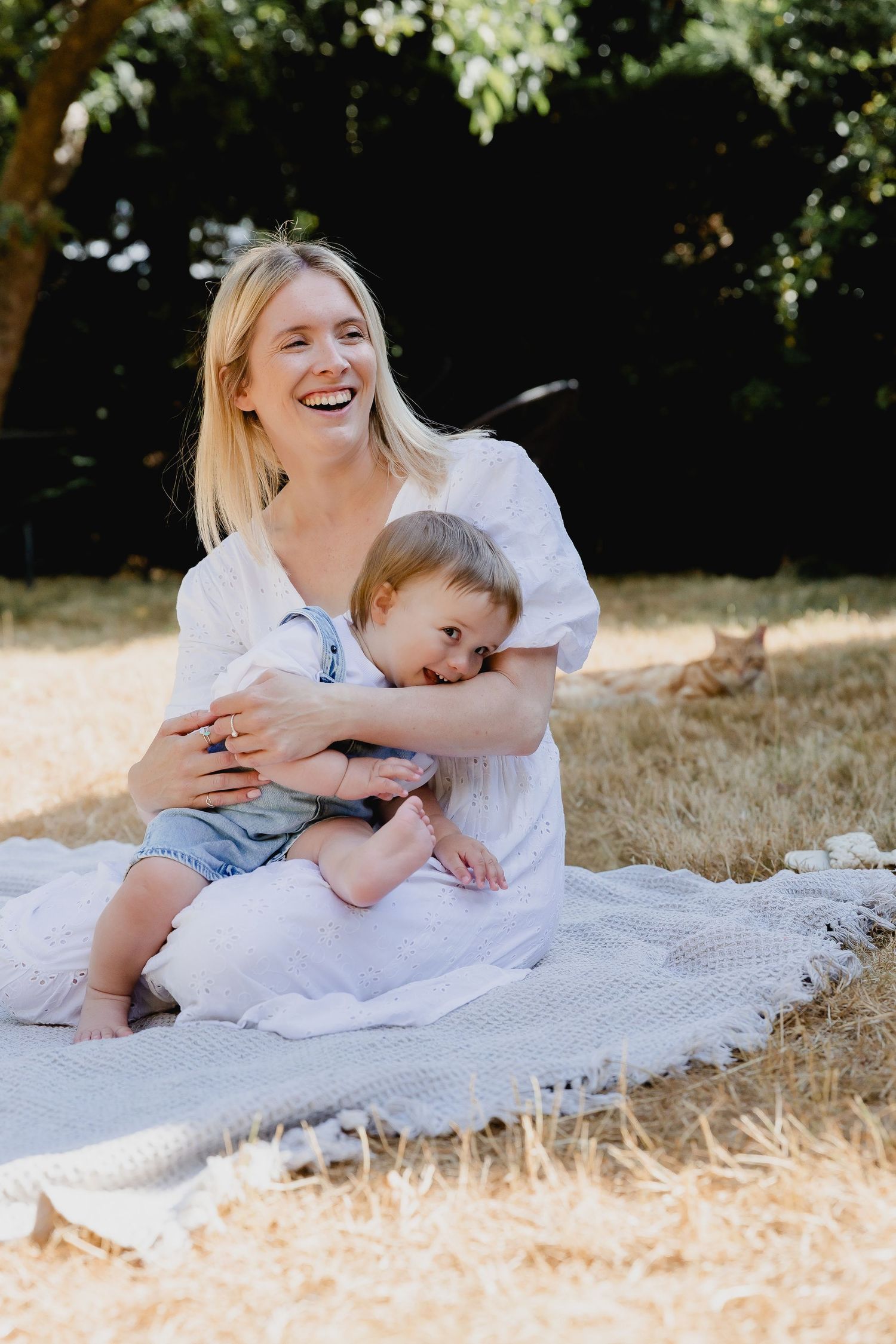 Laughing blonde mother sitting on blanket outdoors hugging her toddler child.