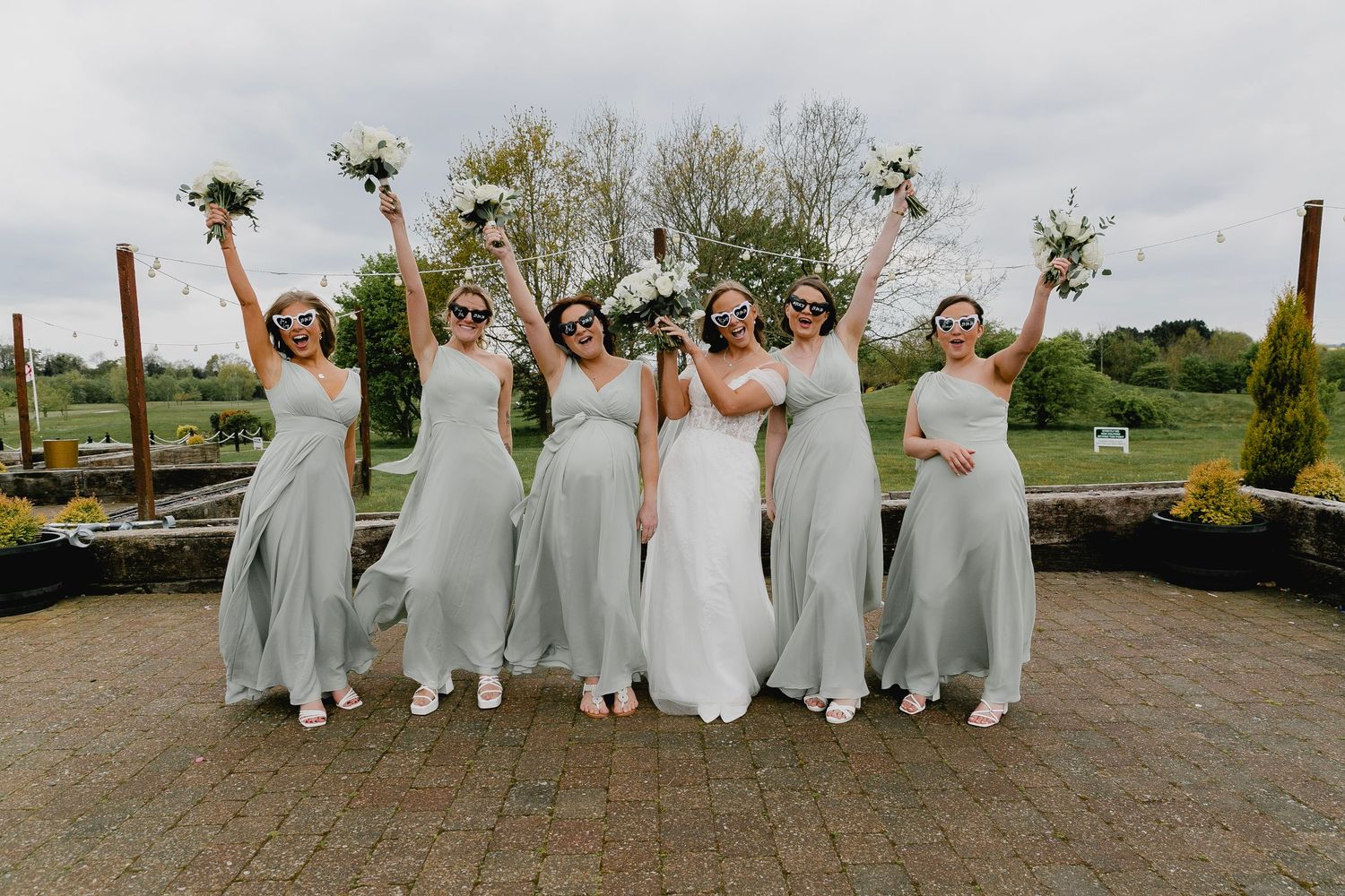 Bride with bridesmaids in sage green dresses celebrating outdoors with bouquets raised.