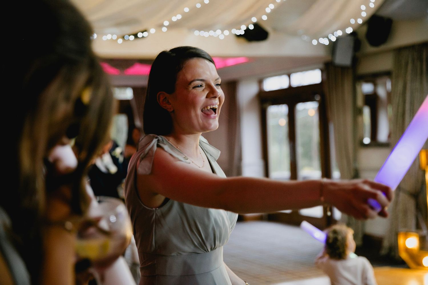 A bridesmaid in a sage green dress laughs joyfully while dancing at a wedding reception with fairy lights.